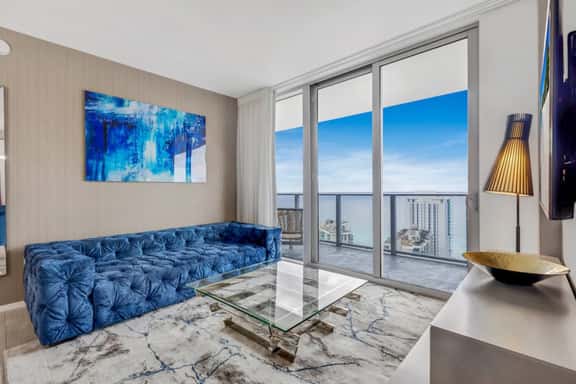 A view of the sparkling ocean sets the scene for this stylish living room featuring a deep blue velvet sofa and a sleek glass coffee table. The large sliding doors open to a balcony, inviting natural light and the refreshing sea breeze inside.